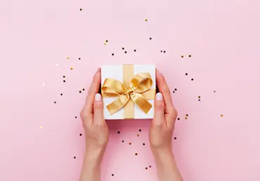 Womans hands holding gift or present box decorated confetti on pink pastel table top view. Flat lay composition for birthday or wedding.