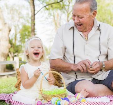 A grandfather looks down at his granddaughter who is smiling and sitting in the grass with an Easter basket. Grandparents can purchase whole life insurance for their grandchildren to help build their financial future.