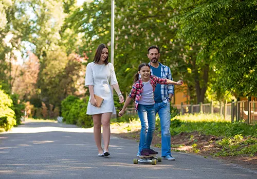 Girl Is Riding A Skateboard In The City Park. Parents Watching Her.
