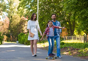 Girl Is Riding A Skateboard In The City Park. Parents Watching Her.