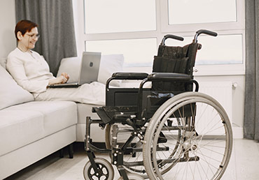 A wheelchair sits in a living room, and a disabled woman sits on a couch with a laptop.