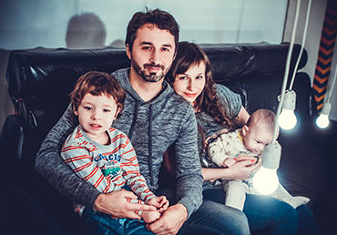 A young family of four sits on a couch for a photo.