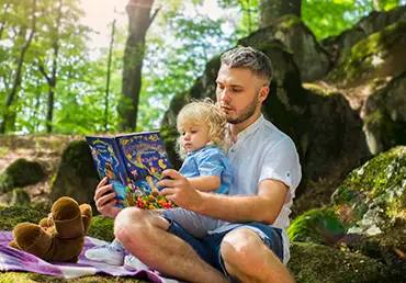 In the forest, a father holds his daughter and reads her a book.
