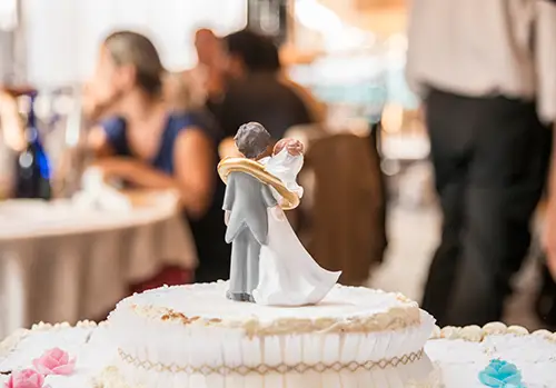 Photo of a husband and wife cake topper in focus on a white wedding cake with a blurry crowd of people in the background.