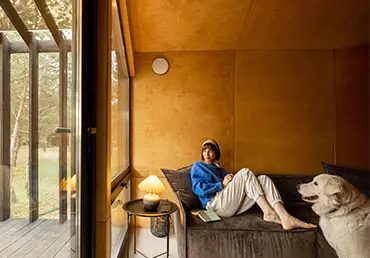 Woman reads a book while sitting relaxed on a couch by the window with her dog at wooden cottage in forest.