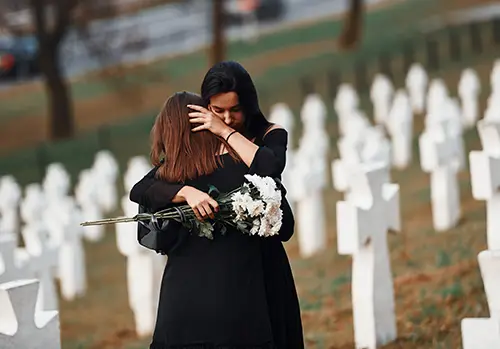 Two young women grieving together in a cemetery with white crosses, representing a funeral and personal loss.